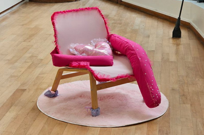 Image of a small pink chair with irregular armrest sizes and a pink heart cushion