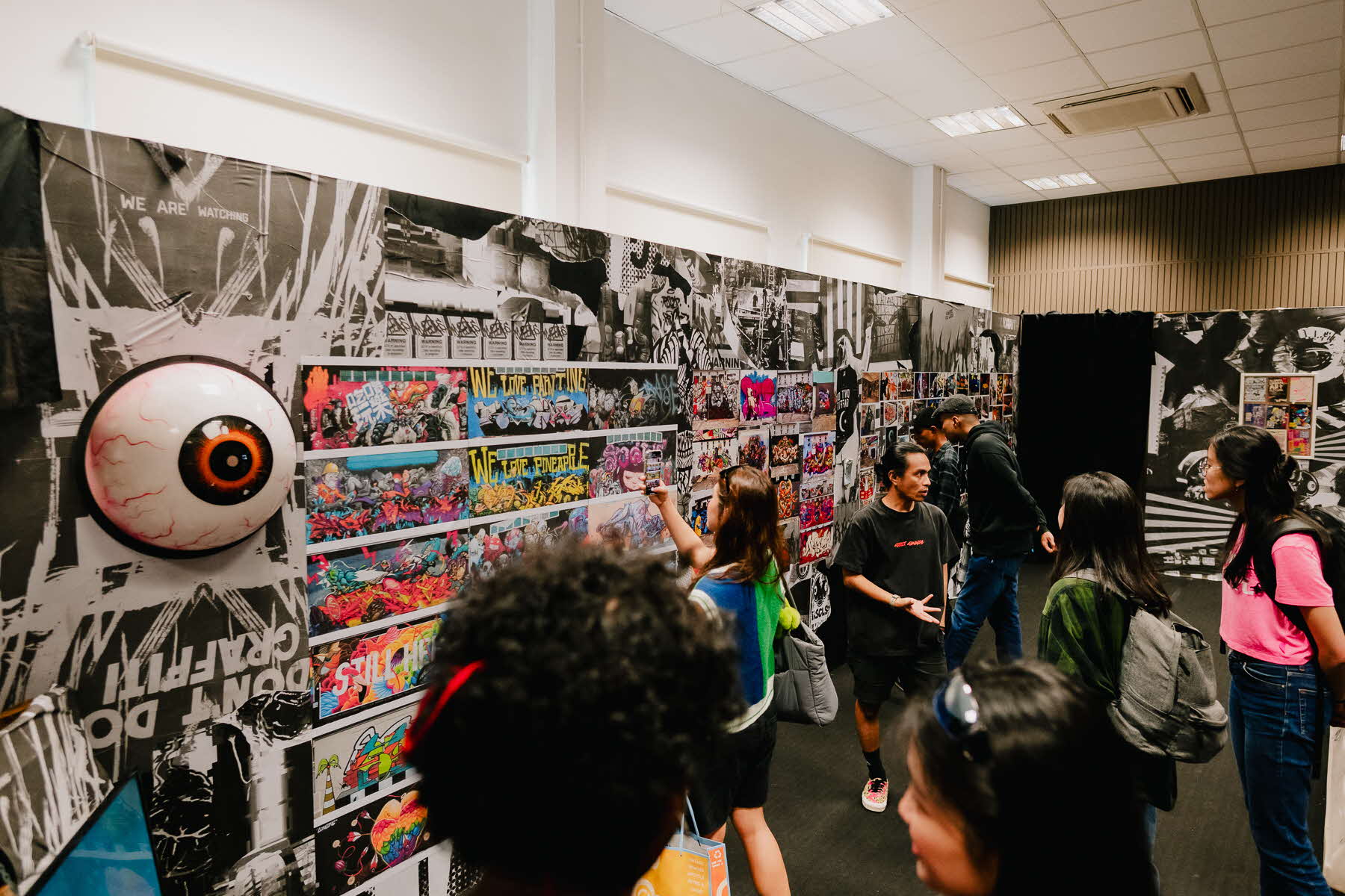 Image of a wall with graffiti and a large eyeball artwork, and people taking pictures of it