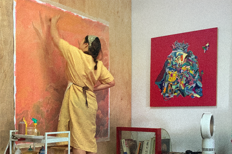 Image of a person in a yellow shirt dress painting on a canvas on a wall. Next to them is a red painting on the other wall.