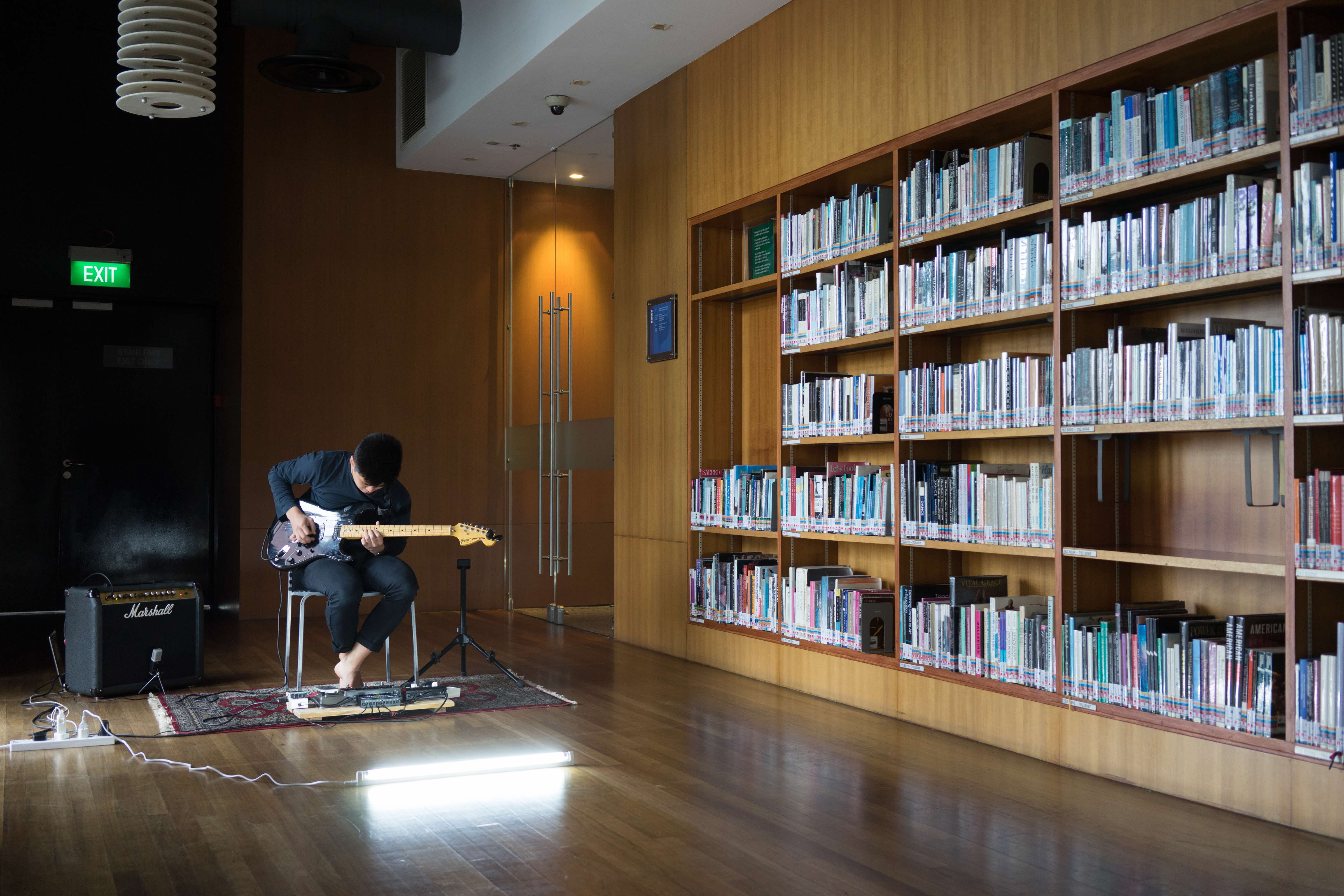 Man playing an electric guitar, with a large LED white light on the floor in front of him, in the now defunct Esplanade Library dance area, which resembles a dance studio, with wooden floors and a bookshelf lined with books.