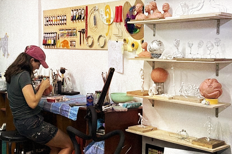 Image of a person in a black tshirt and shorts and maroon cap working on a detailed artwork, surrounded by shelves and a wall of sculptures