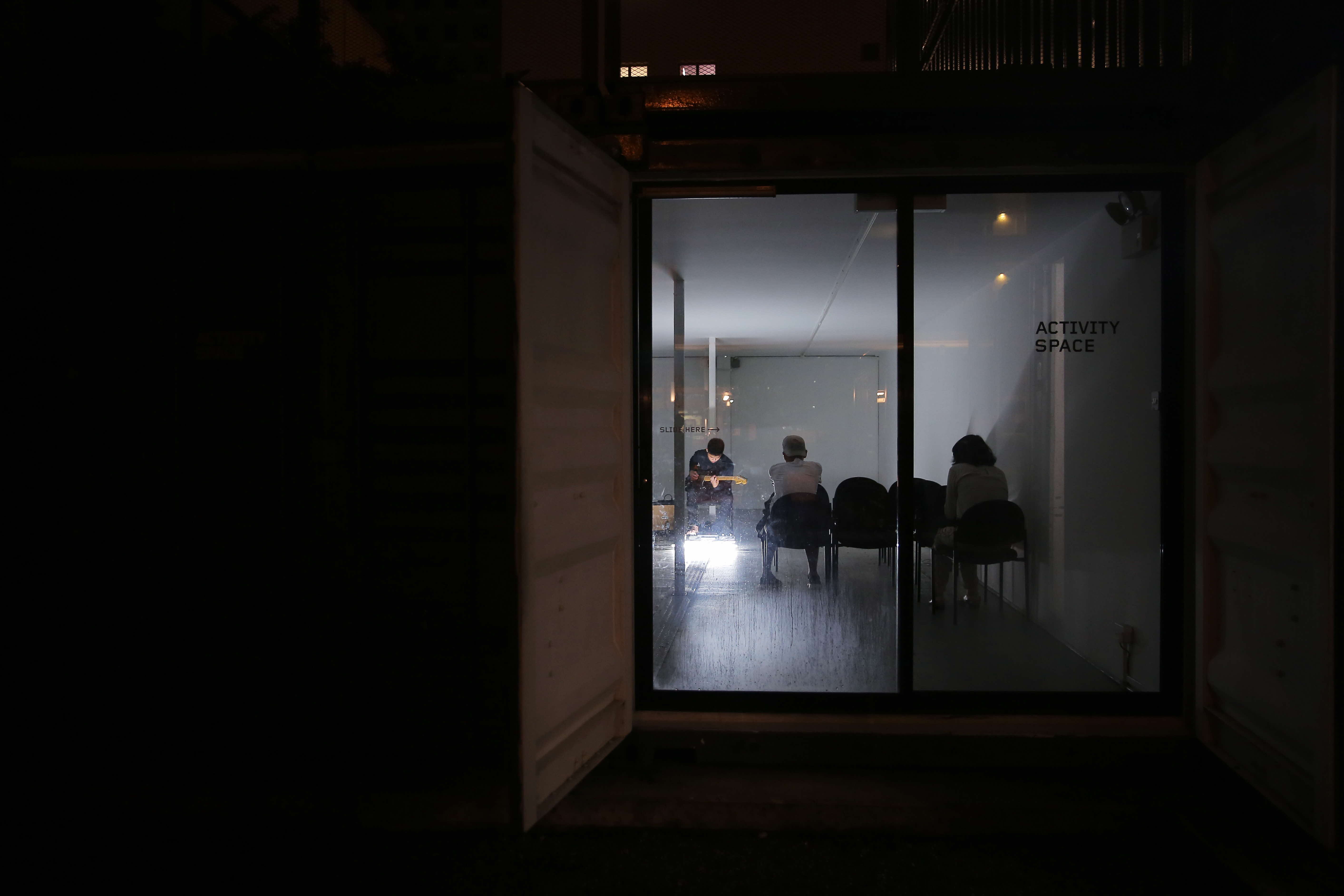 Dark space illuminated only by a room with a glass door, which has a solitary white light, and audiences can be seen listening to a musician playing an instrument.