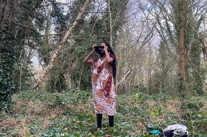 Image of a person in a red and white patterned dress holding a pair of binoculars, surrounded by a forest clearing