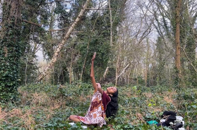 Image of a person in a red and white patterned dress with arms raised asymmetrically up to the sky, surrounded by a forest clearing