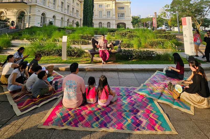 Image of audience in public sitting on colourful batik mats listening to a storyteller